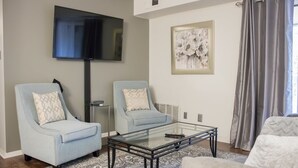 A living room with two light blue armchairs, a glass coffee table, a flat-screen TV mounted on the wall, and a framed abstract painting. - Modern Upscale Private Room (Raleigh)
