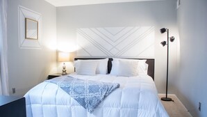 A neatly made bed with a patterned throw, a bedside lamp, and a floor lamp in a modern bedroom with a geometric wall design. - Modern Upscale Private Room (Raleigh)