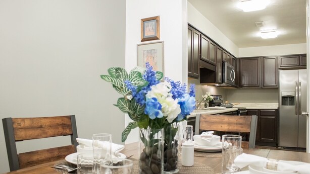 A dining area with a wooden table set for four, surrounded by dark wooden chairs. A vase with blue and white flowers is placed on the table. - Modern Upscale Private Room (Raleigh)