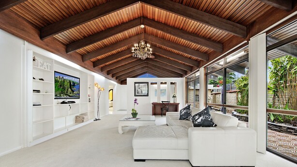 A spacious living room with a high ceiling, white sofas, a flat-screen TV, and a large window offering a view of the outdoors.