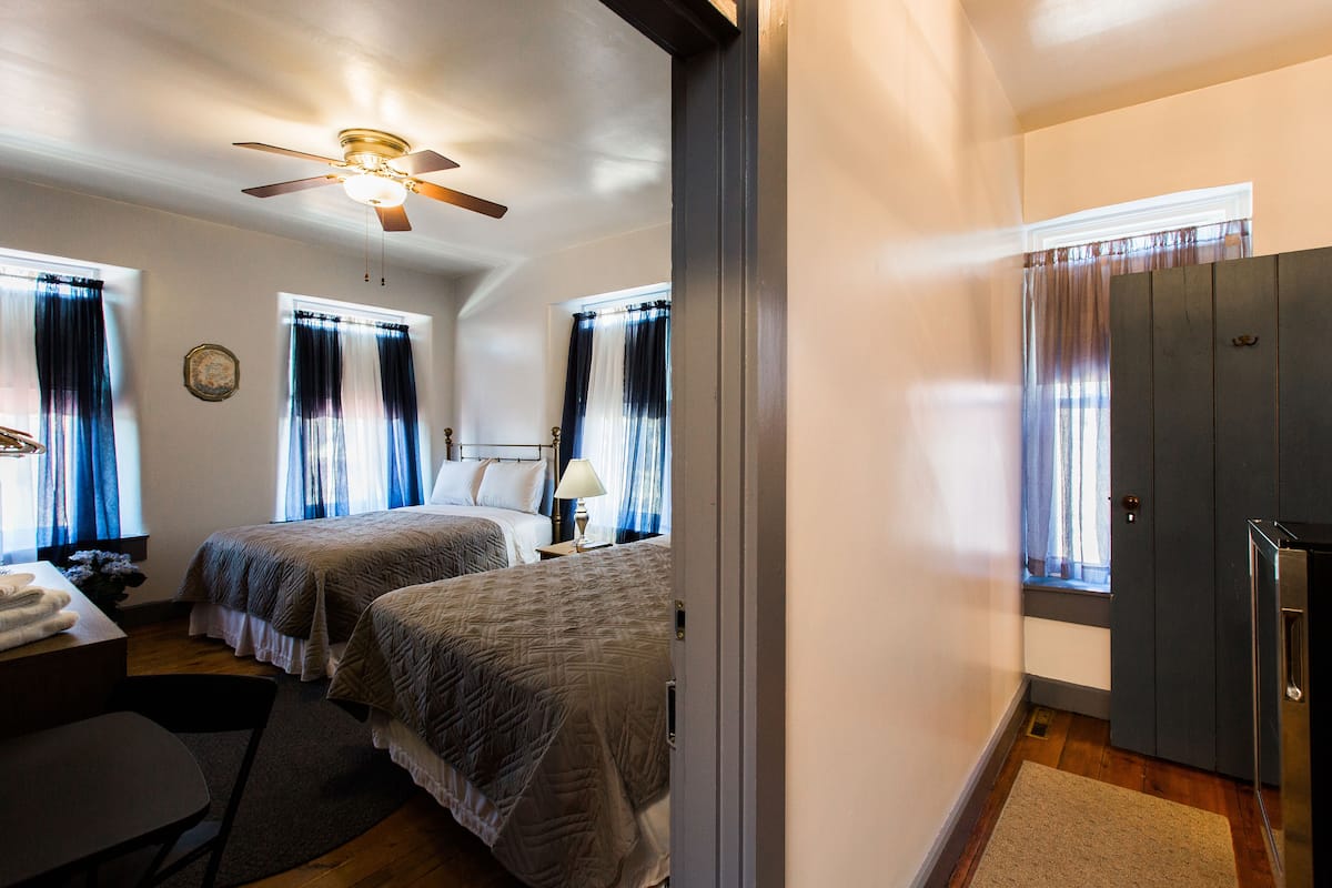 Une chambre d’hôtel avec deux lits, un bureau, une chaise, un ventilateur de plafond et une fenêtre avec des rideaux.