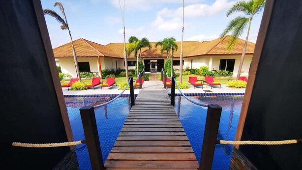 A wooden deck leading to a pool with red lounge chairs and palm trees in the background. - Tamara Resort Bangsary (Sattahip)