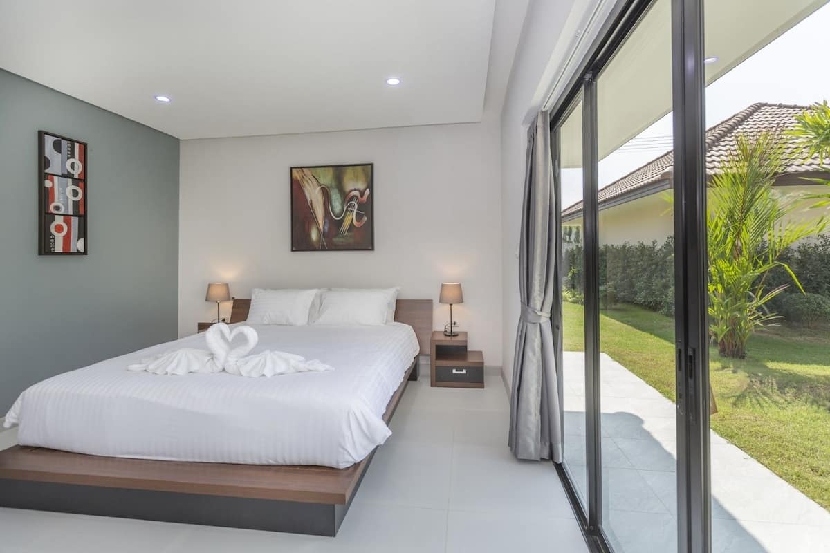 Quarto moderno com uma cama grande, duas mesas de cabeceira com abajures, um quadro na parede e vista para um jardim através de uma porta de vidro deslizante.