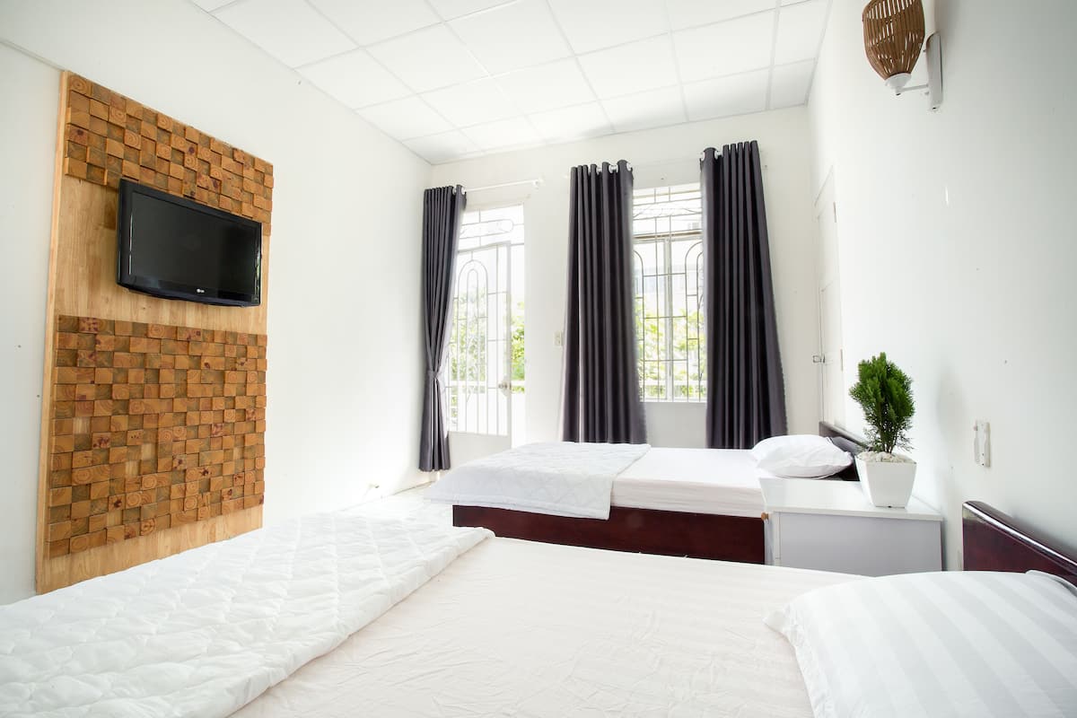 Habitación de hotel con cabecera de madera, televisor de pantalla plana integrado, cama con ropa de cama blanca, mesita de noche con una planta y una ventana con cortinas.