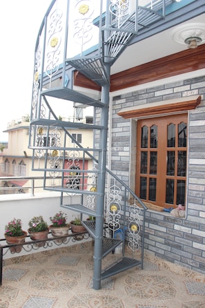 Un balcone esterno su più livelli con una scala in metallo, piante in vaso e una finestra con cornice in legno.
