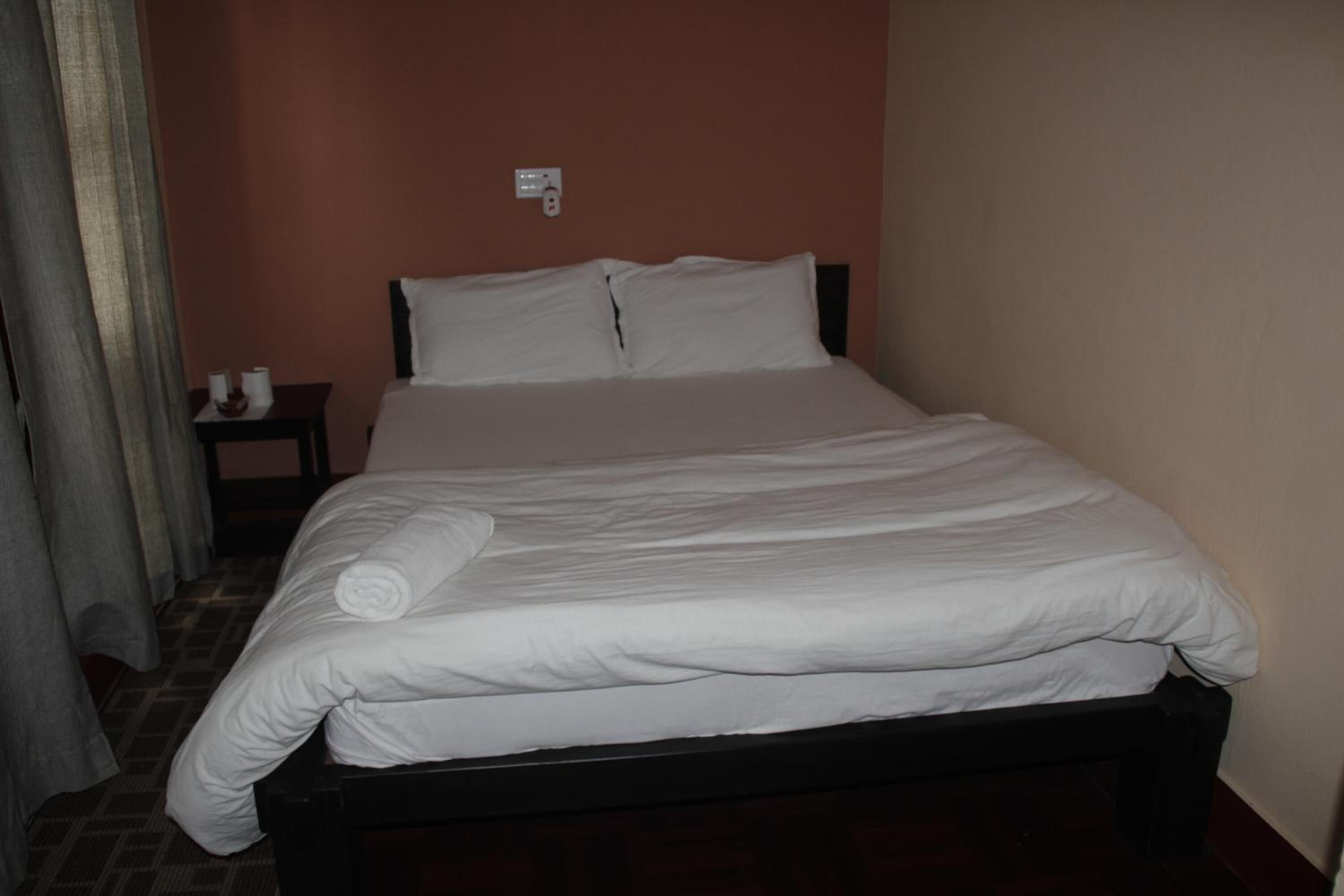 Una cama bien tendida con sábanas blancas, un cabecero de madera oscura y una mesita de noche con una lámpara y una taza.