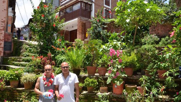 Ein Garten mit Blumentöpfen und Blumen, ein Backsteinhaus und eine Person, die eine Blume hält.