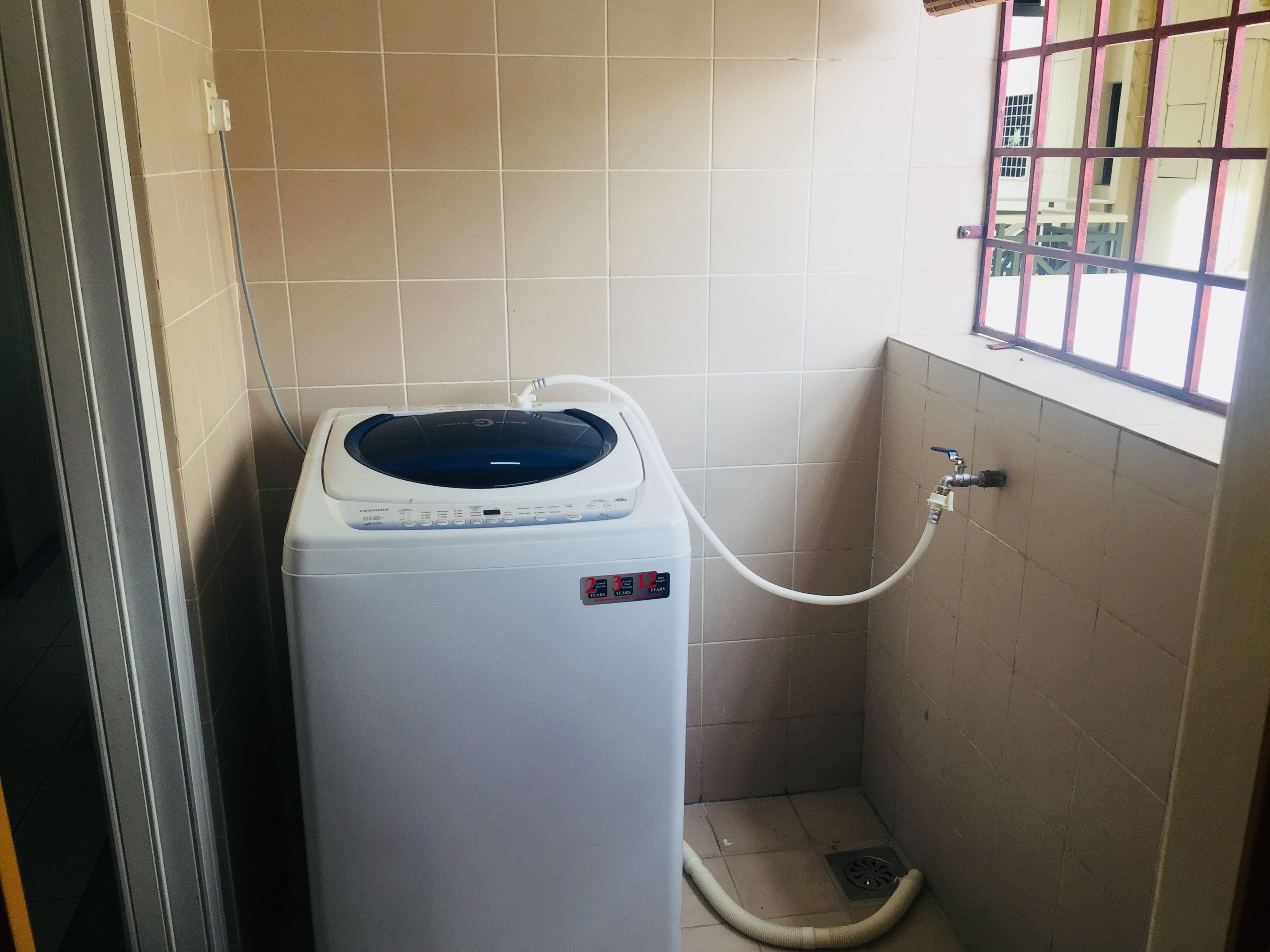 Une petite machine à laver blanche à chargement frontal, installée dans une petite salle de bain carrelée, avec une fenêtre et une grille métallique.