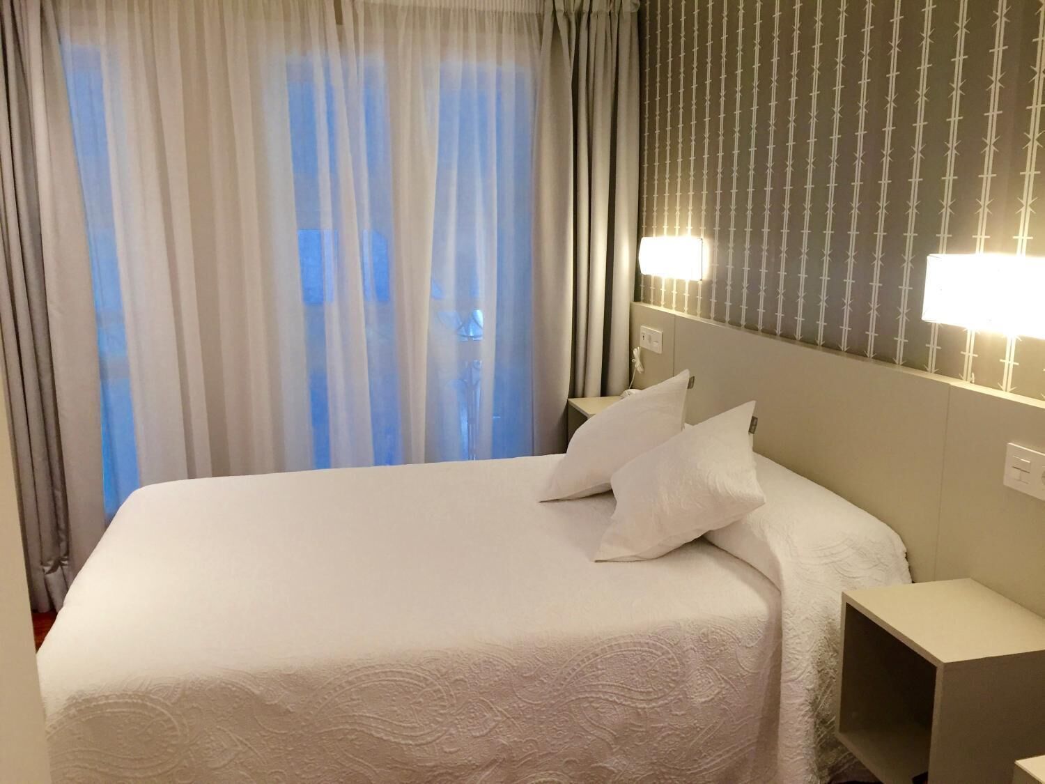 Una habitación de hotel con una cama grande, ropa de cama blanca, una mesita de noche y una lámpara fijada a la pared.