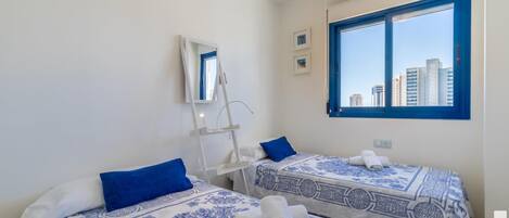 Ett hotellrum med två sängar, var och en med ett blått och vitt mönstrat överkast, en blå kudde och hoprullade handdukar. Det finns ett fönster med en blå ram och en spegel på väggen.