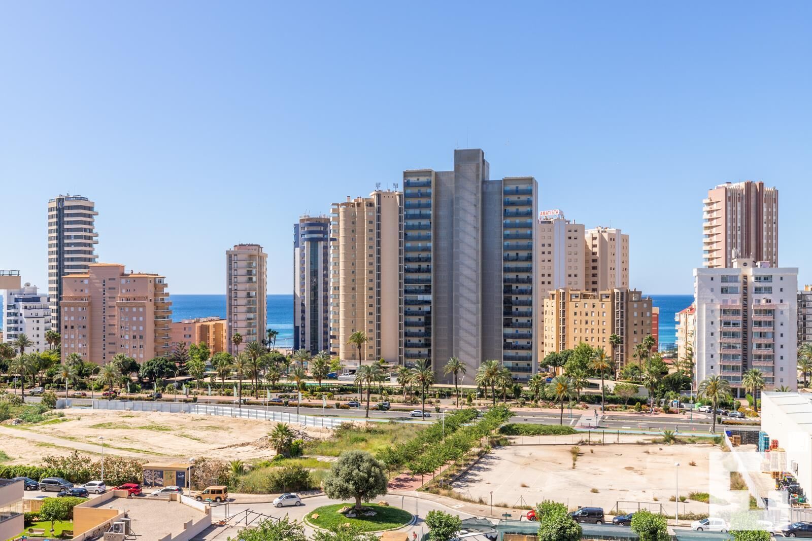 A coastal cityscape with high-rise buildings, a clear sky, and a sandy area with palm trees.