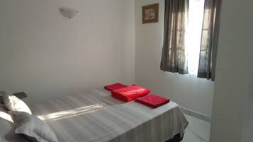 Une petite chambre avec un lit simple, un mur blanc uni, un petit tableau et une fenêtre avec des rideaux.
