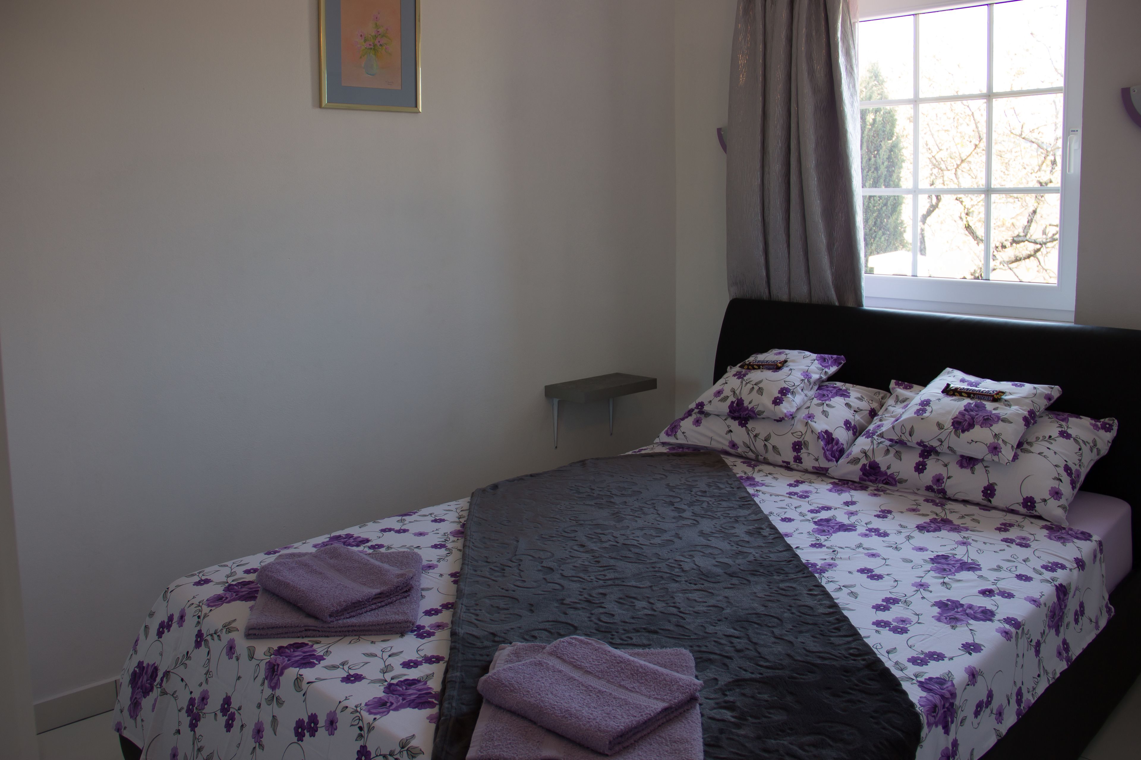Una cama bien hecha con ropa de cama floral y un cabecero oscuro, una ventana con cortinas y una mesita de noche pequeña.
