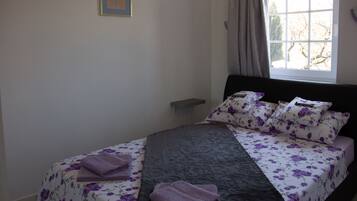 Una cama bien hecha con ropa de cama floral y un cabecero oscuro, una ventana con cortinas y una mesita de noche pequeña.