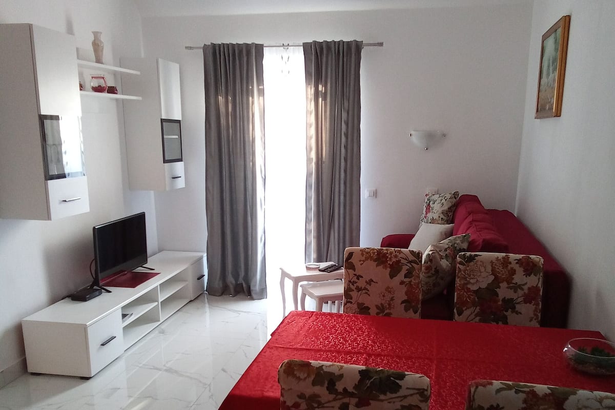 A modern living room with a dining area, white walls, a flat-screen TV, a red dining table, floral chairs, and a large window with curtains.