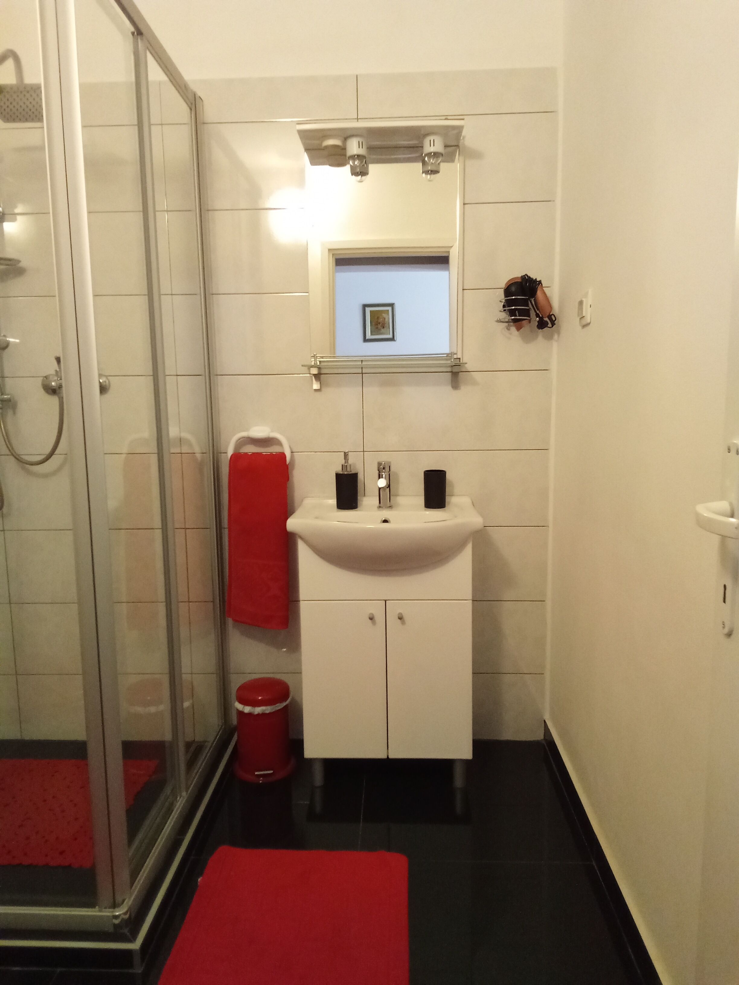 Um banheiro com box de vidro, pia branca com dois dosadores de sabonete pretos e uma toalha vermelha pendurada na parede.