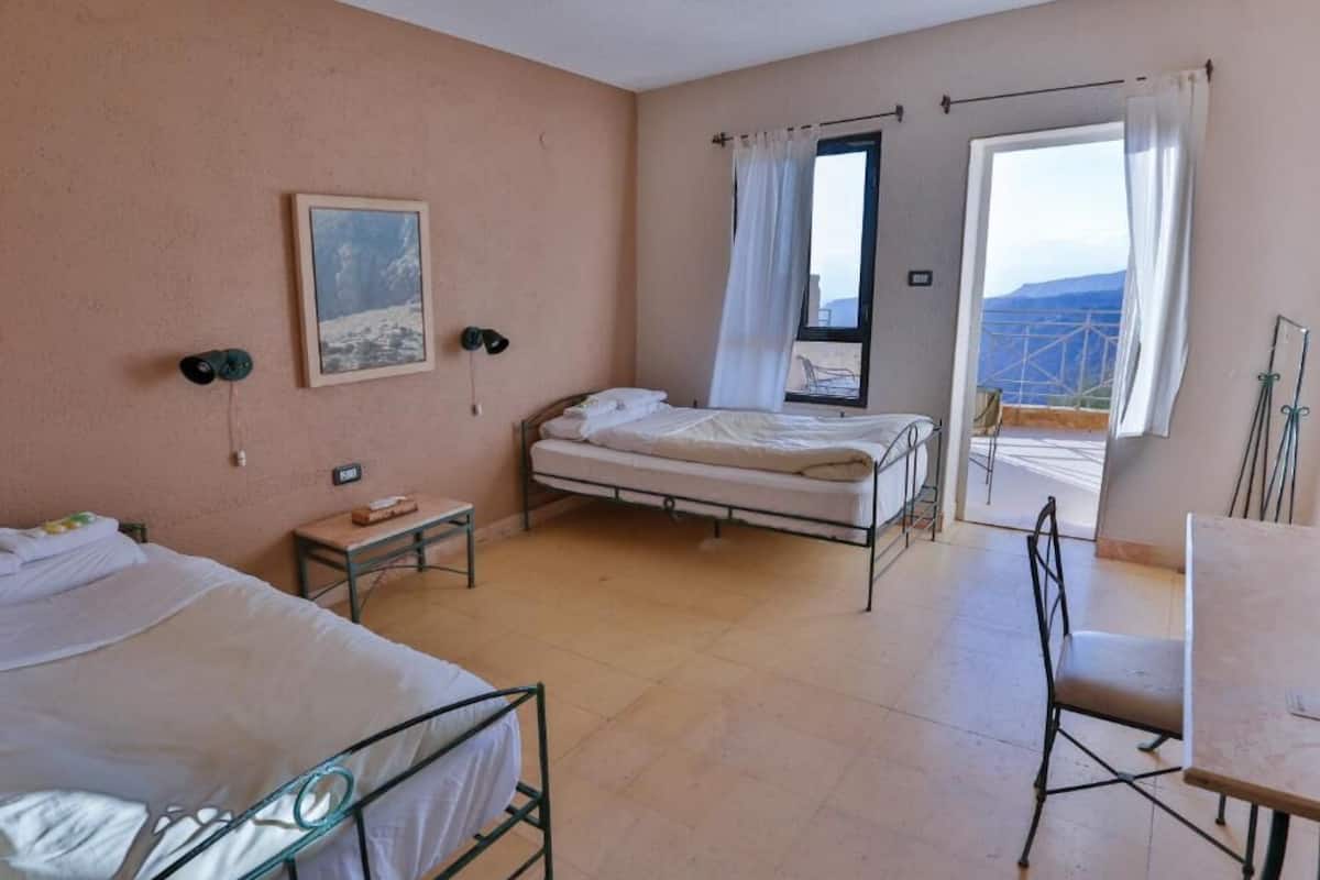 A bedroom with two beds, a desk, and a chair. There is a window with a view of the mountains.