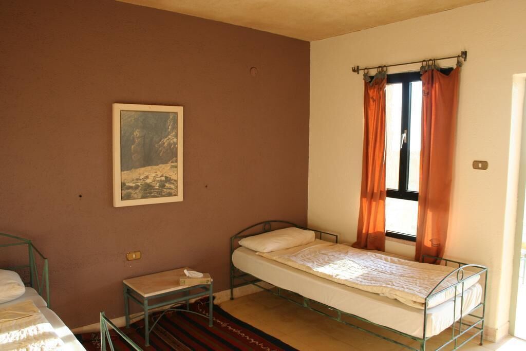 Een kleine kamer met een enkel bed met metalen frame, een klein tafeltje, een ingelijst foto aan de muur en een raam met oranje gordijnen.
