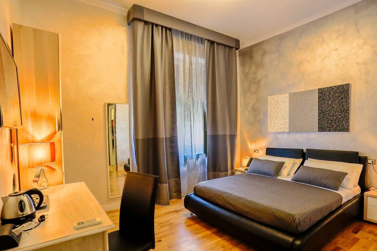 A modern hotel room with a large bed, a desk with a kettle, and a chair.