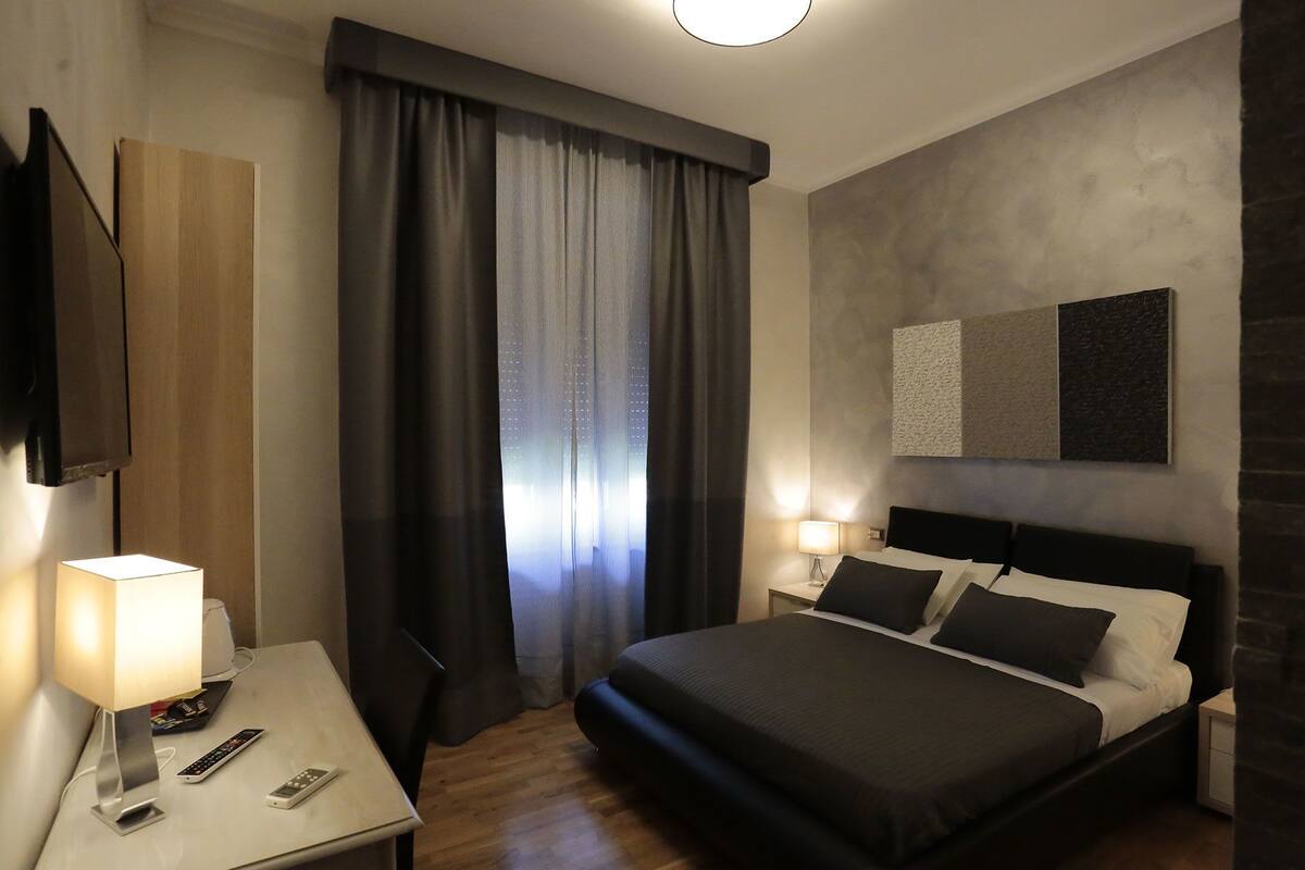A modern hotel room with a large bed, bedside lamps, a flat-screen TV, and a large window with curtains.