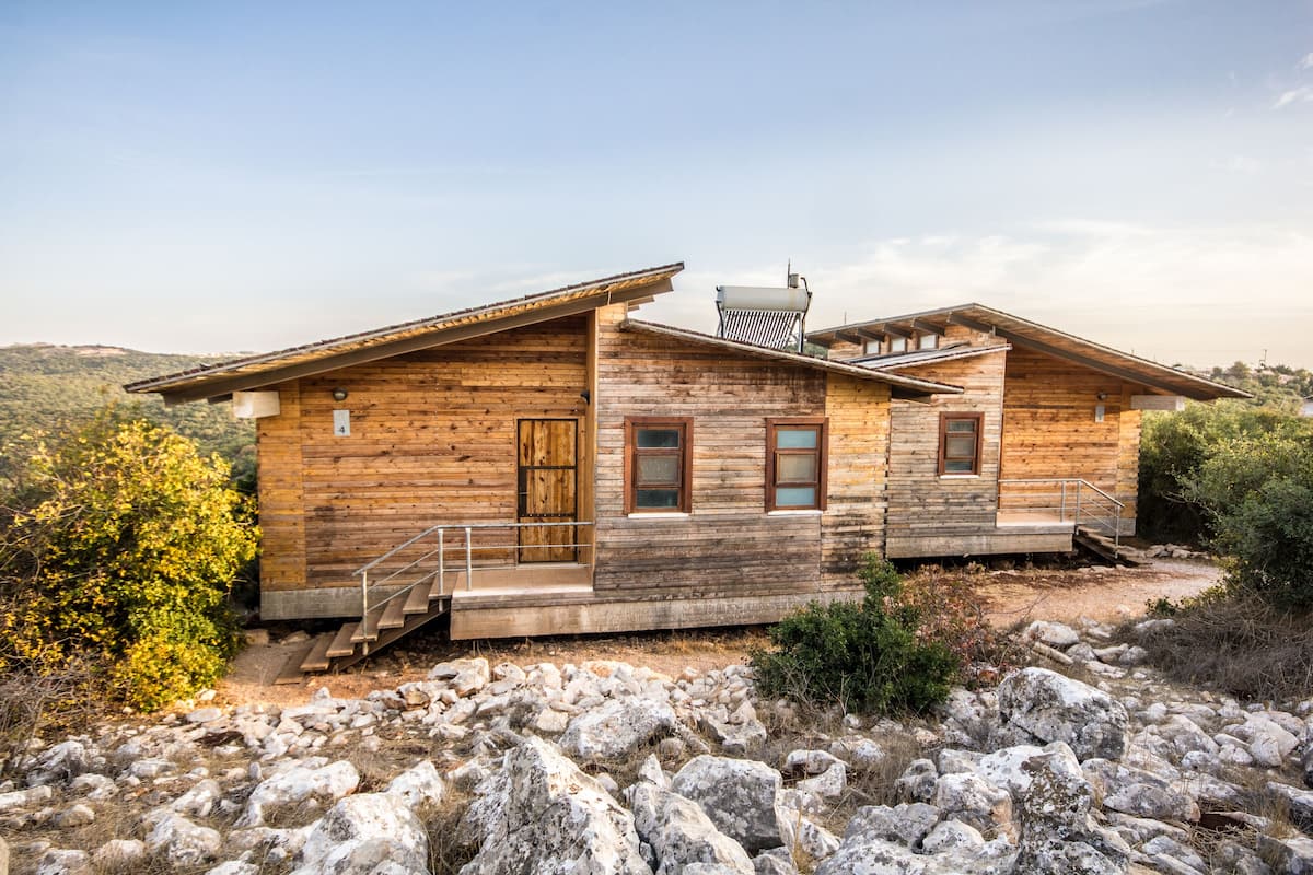 Une cabane en bois avec un escalier menant à une porte en bois, située sur un terrain rocheux et offrant une vue sur des collines en arrière-plan.
