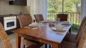 A dining area with a wooden table set for six, surrounded by wicker chairs, a kitchen with dark cabinets and a white oven, and a balcony with a view of trees. - MONTAUROUX 30MN OF CANNES APARTMENT CONDITIONING TERRACE GARDEN PRIVATE POOL (Montauroux)