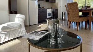 A modern living room with a glass table displaying wine glasses and a magazine, a white sofa, a wicker chair, and a kitchen in the background. - MONTAUROUX 30MN OF CANNES APARTMENT CONDITIONING TERRACE GARDEN PRIVATE POOL (Montauroux)