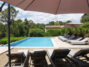 Une piscine entourée de verdure, dotée de chaises longues et de parasols, et surmontée d’une structure à toit carrelé.