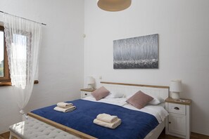 Une chambre d’hôtel comprenant un lit, deux tables de chevet, une lampe, une œuvre d’art accrochée au mur et une fenêtre avec des rideaux légers.
