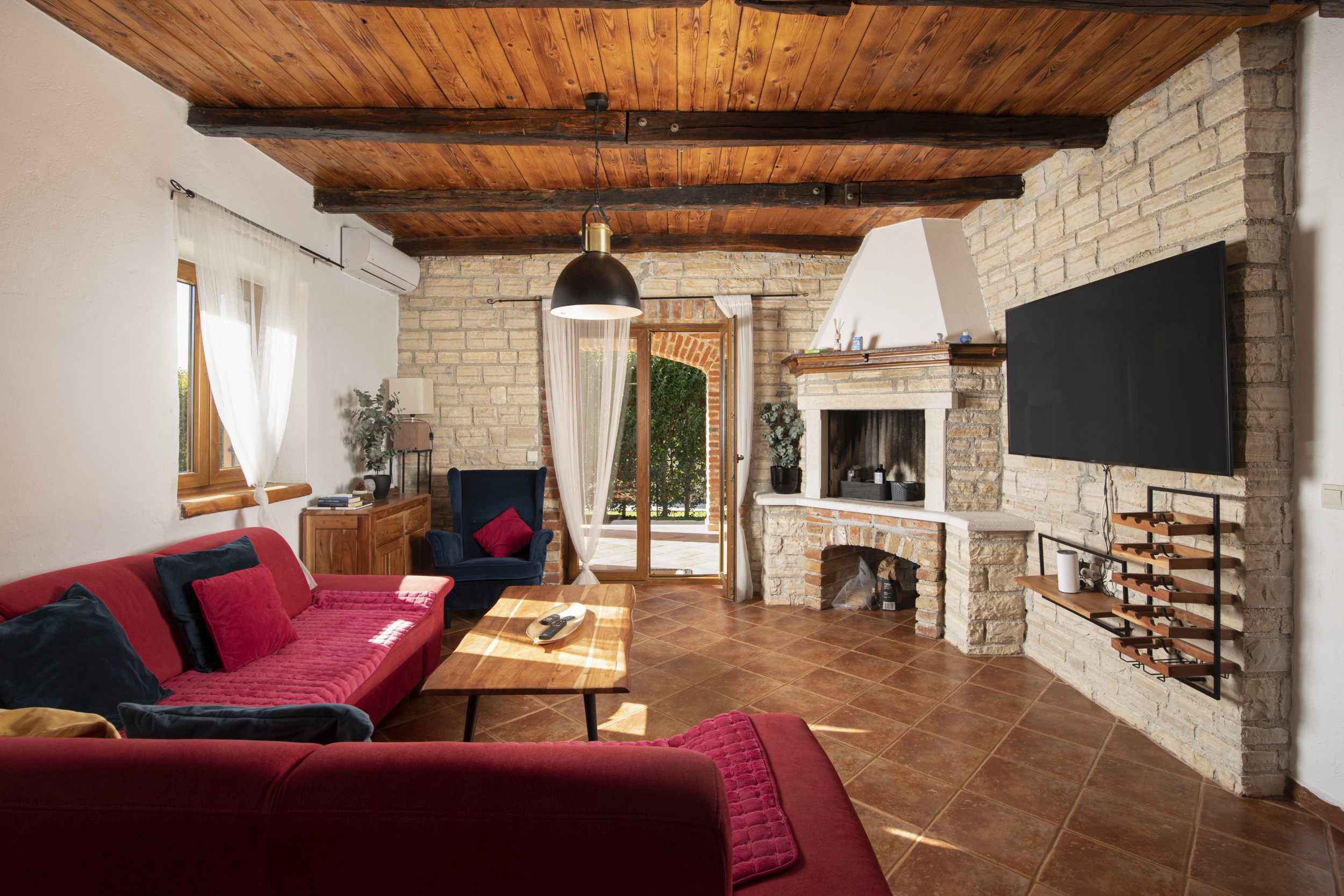 A cozy living room with a red sofa, a wooden ceiling, a stone fireplace, and a TV mounted on the wall.