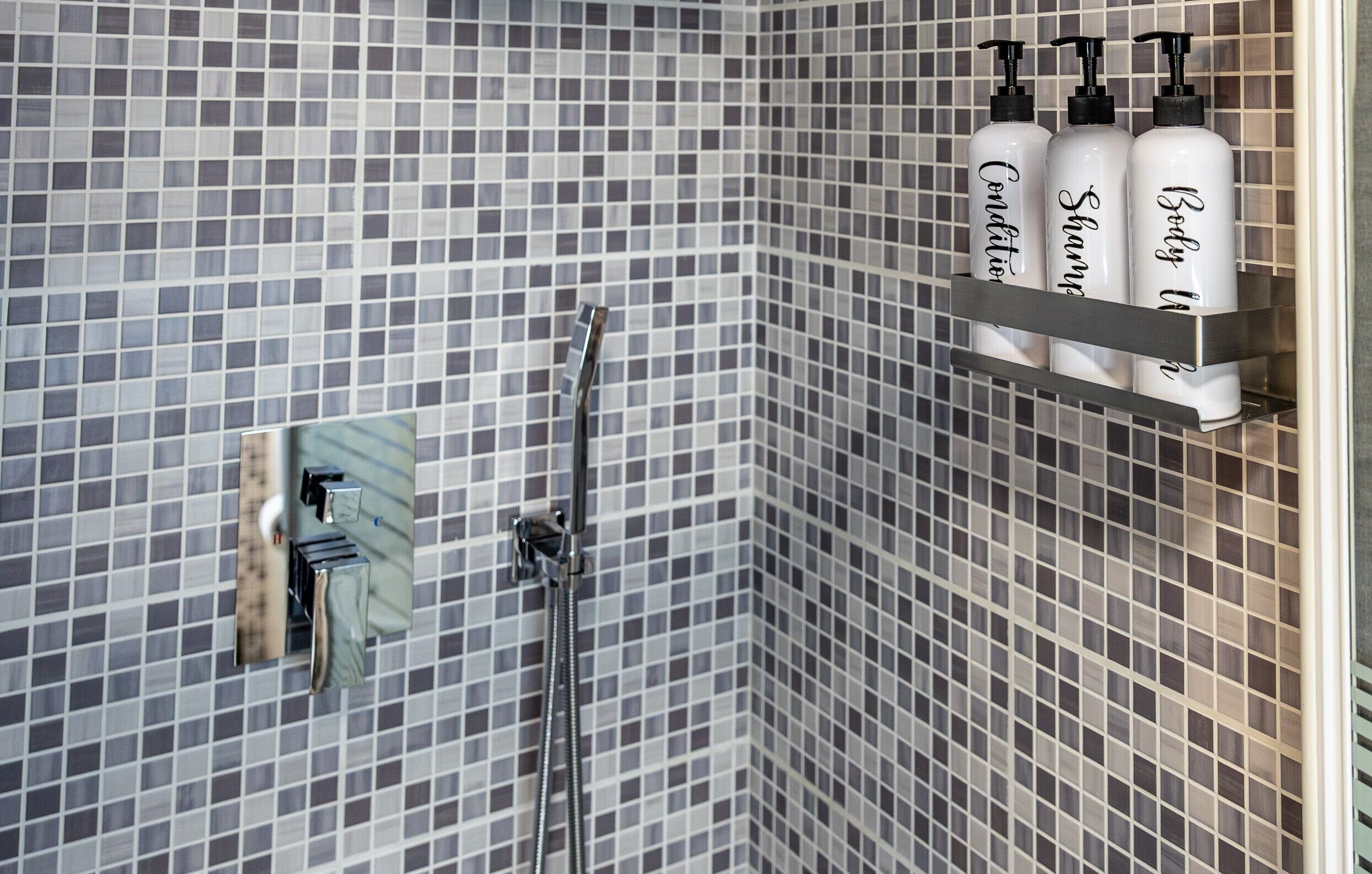 Une salle de bain avec un mur de douche recouvert de carreaux, une douchette à main et une étagère sur laquelle sont posées trois bouteilles étiquetées « Après-shampoing », « Shampoing » et « Gel douche ».