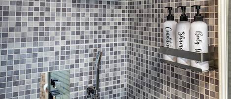 Une salle de bain avec un mur de douche recouvert de carreaux, une douchette à main et une étagère sur laquelle sont posées trois bouteilles étiquetées « Après-shampoing », « Shampoing » et « Gel douche ».
