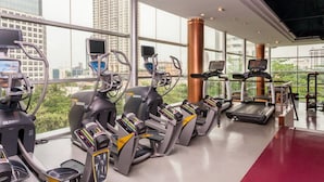Ein Fitnessstudio mit Laufbändern, Crosstrainern und stationären Fahrrädern, großen Fenstern und einem roten Boden.