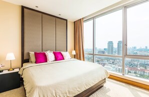 Una habitación de hotel con una cama grande, una mesita de noche, una lámpara y vistas a la ciudad a través de un ventanal amplio.