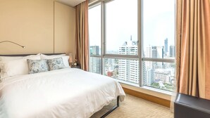 Ein Hotelzimmer mit einem großen Bett, einem Nachttisch, einer Lampe und Blick auf die Stadt durch große Fenster.