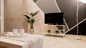 Un salon moderne avec un téléviseur à écran plat posé sur un meuble blanc, une plante en pot et une table basse recouverte d’une nappe blanche.