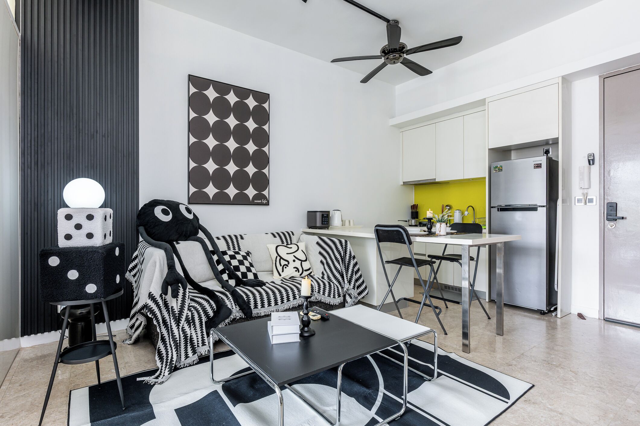 Una sala moderna con un sofá blanco y negro, una mesa de comedor y una zona de cocina con refrigerador y microondas.