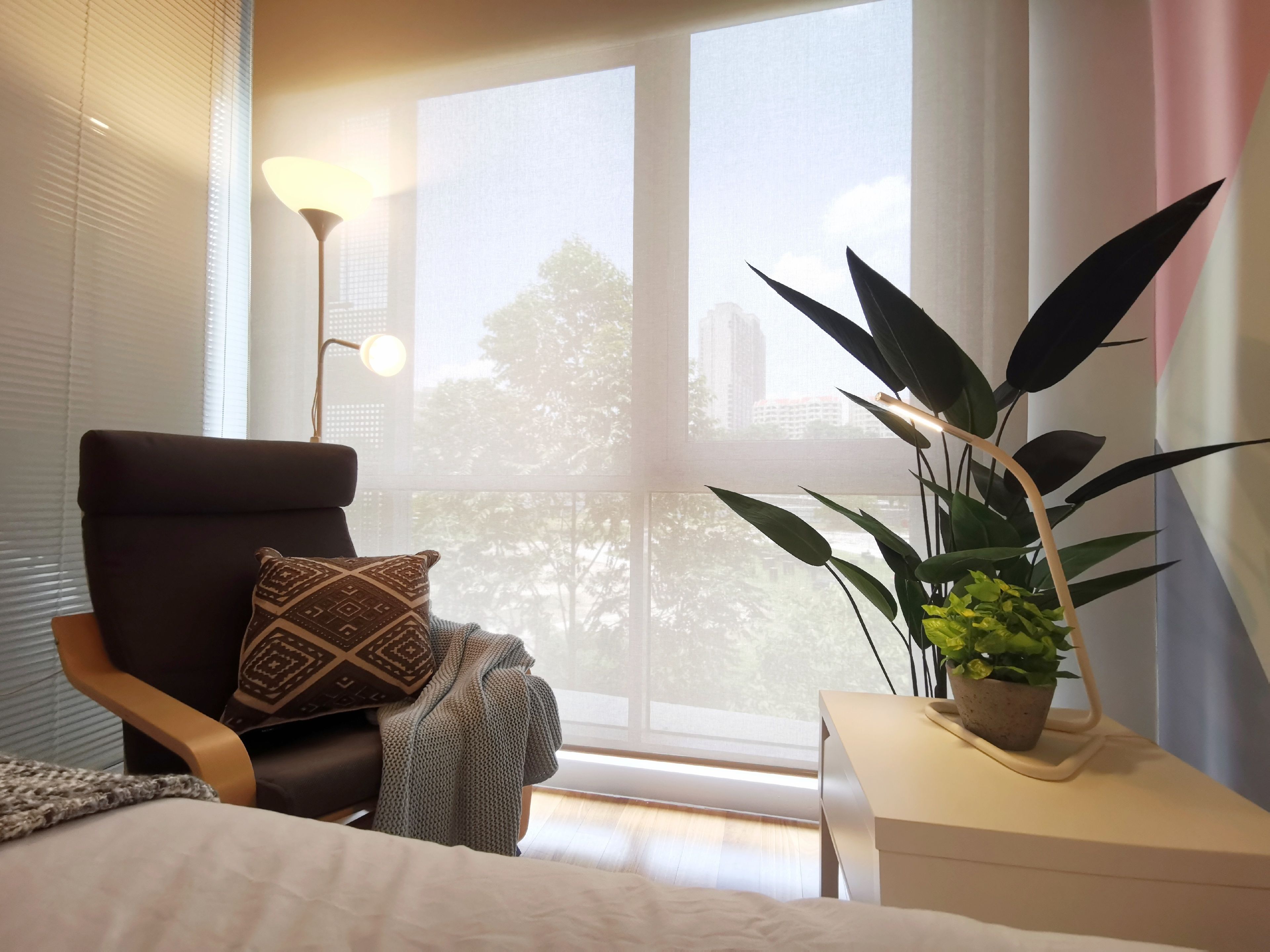 Een moderne slaapkamer met een groot raam, een comfortabele stoel, een nachtkastje met een plant en een lamp.