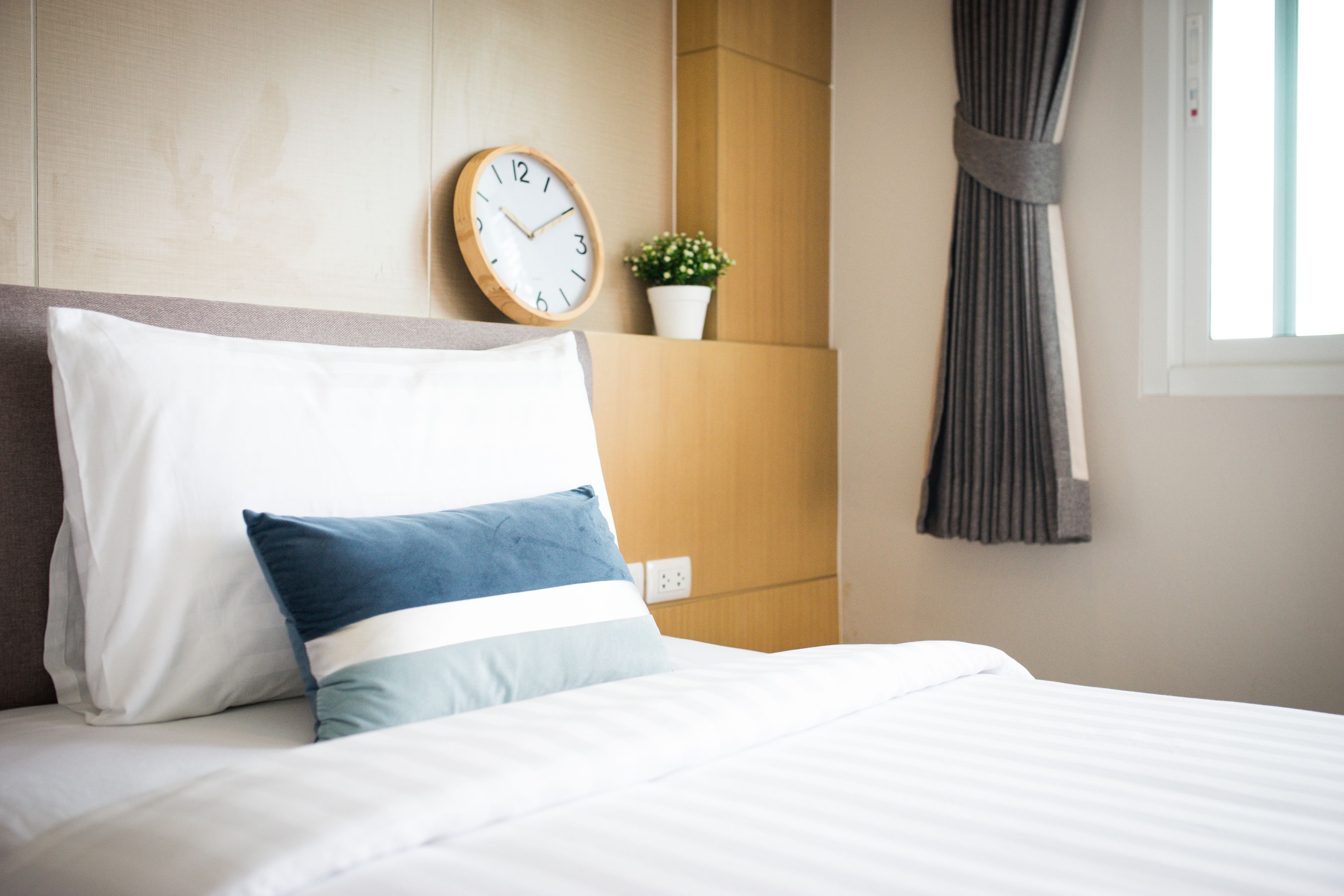 Una cama bien hecha con almohadas blancas y azules, una mesita de noche de madera con un reloj y una pequeña planta, y una ventana con cortinas.