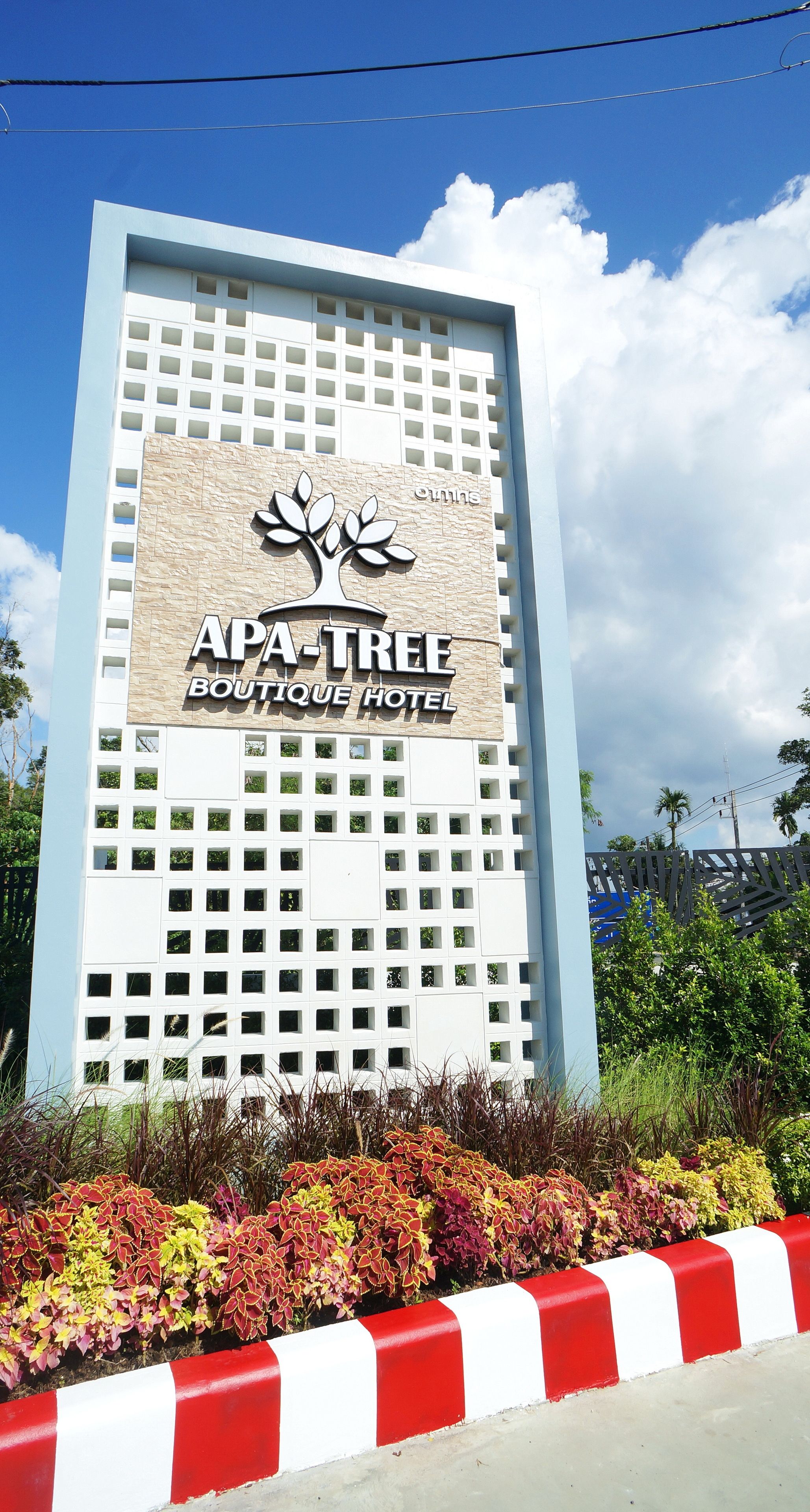 Letrero del APA-TREE Boutique Hotel con un logo de árbol, rodeado de plantas coloridas y una bordeadora roja y blanca.