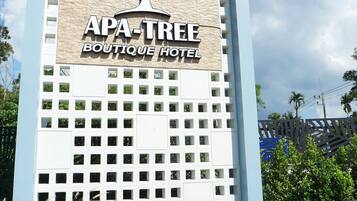 Letrero del APA-TREE Boutique Hotel con un logo de árbol, rodeado de plantas coloridas y una bordeadora roja y blanca.