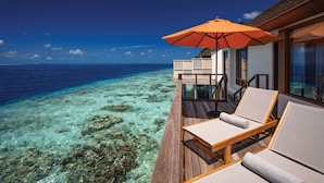 Una terraza de madera con dos sillones de descanso y una sombrilla naranja, con vista a un mar azul cristalino y un arrecife de coral.