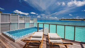 Una terraza de madera con piscina y sillas de descanso, con vistas a aguas turquesas y un cielo despejado.