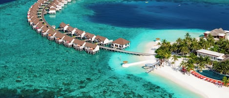 An aerial view of a resort with overwater bungalows connected by walkways, a beach area with palm trees, and clear turquoise waters.