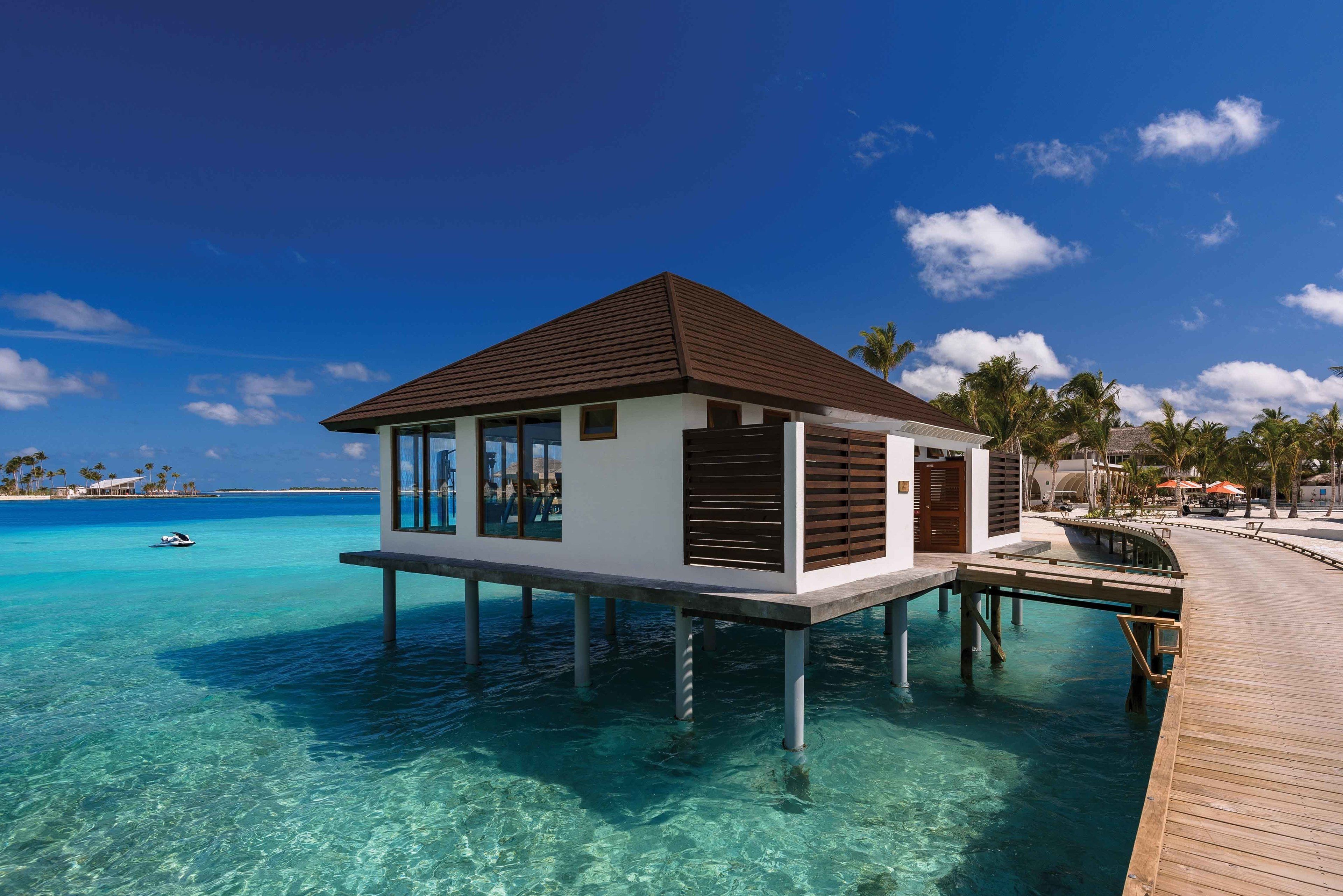 An overwater bungalow with a wooden deck, clear turquoise water, and a view of the ocean and palm trees.