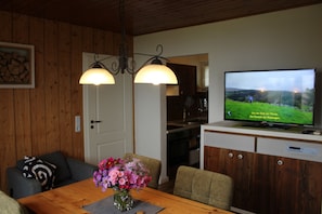Un área de comedor con una mesa de madera, sillas y un jarrón de flores. Un televisor está montado en un mueble de madera sobre la mesa.