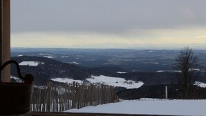 A view through a window with a wooden fence and a metal bucket in the foreground, overlooking a snowy landscape with distant mountains. - Newly renovated holiday home in the Bavarian Forest (Schöfweg)