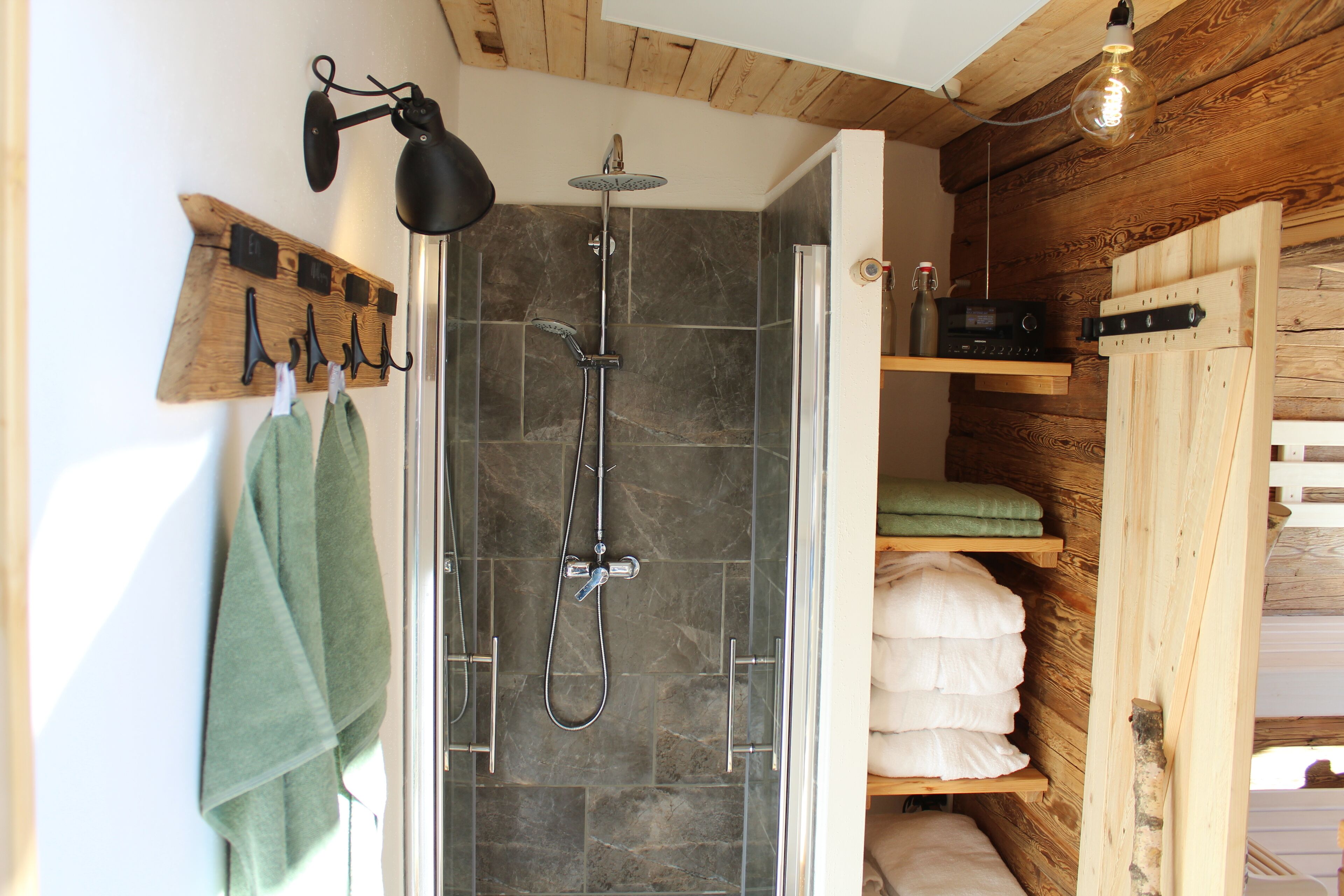 Une douche avec une porte en verre, une étagère en bois où sont posées des serviettes et un porte-serviettes fixé au mur.