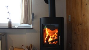 A wood-burning stove with visible flames, a window with curtains, and a wooden wall. - Newly renovated holiday home in the Bavarian Forest (Schöfweg)