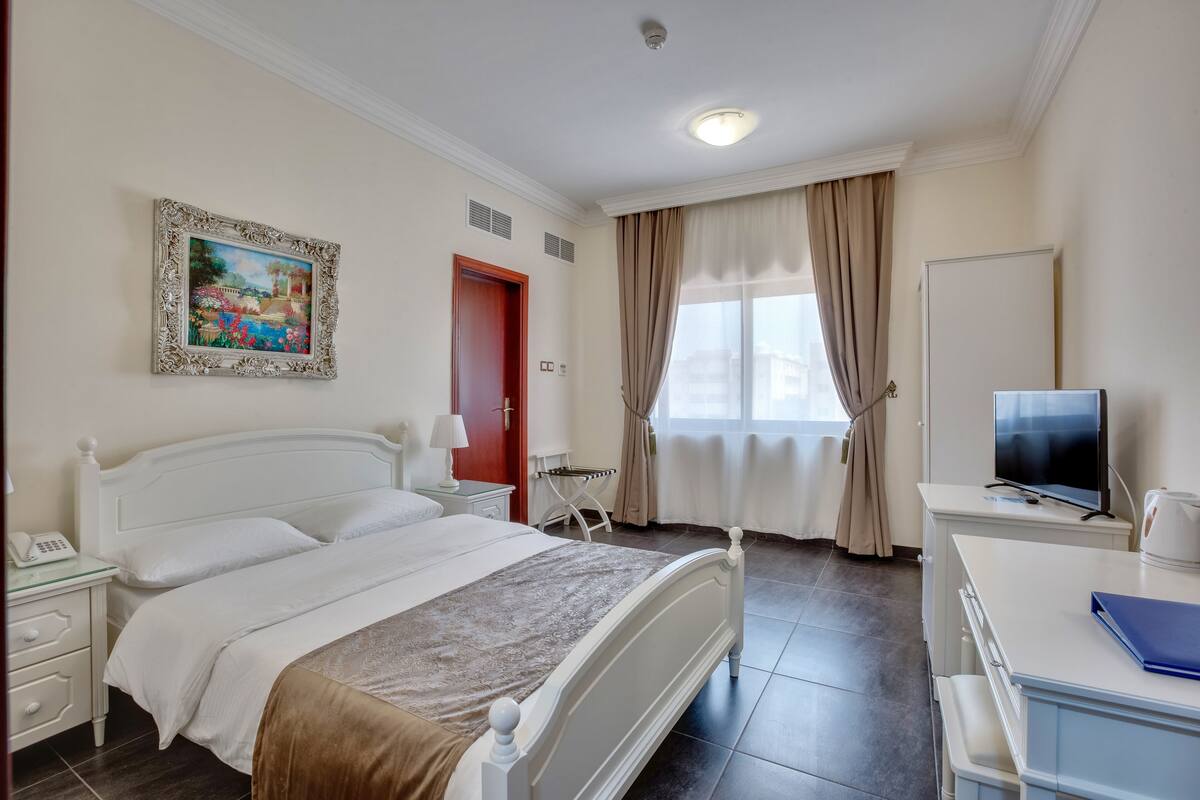 A hotel room with a bed, bedside table, lamp, TV, and a painting on the wall.
